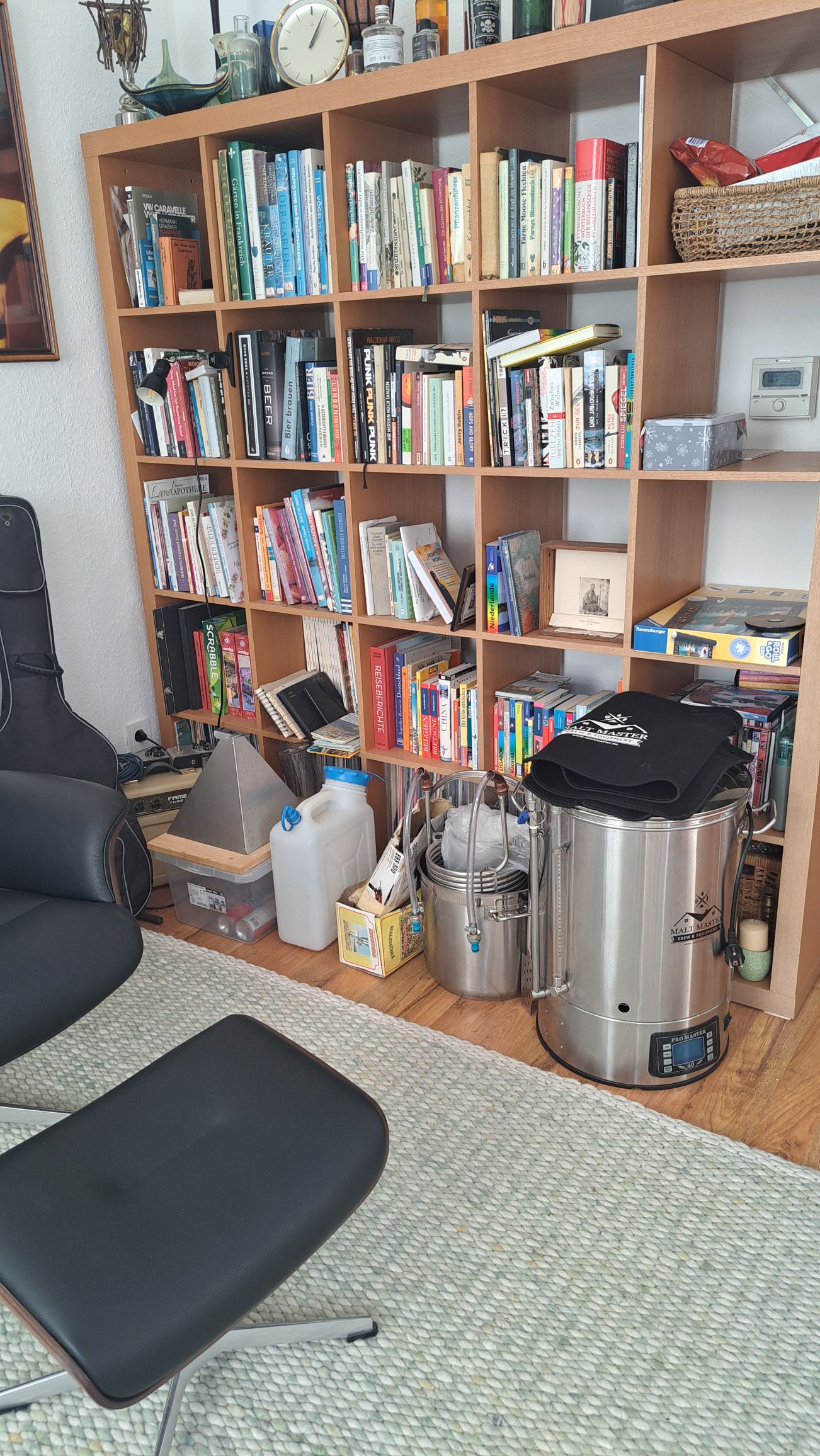 Verschiedene Brauutensilien wie Malzmühle, Wasserkannister, Braukessel, Nachgusstopf) aufgereiht vor einem Bücherregal.